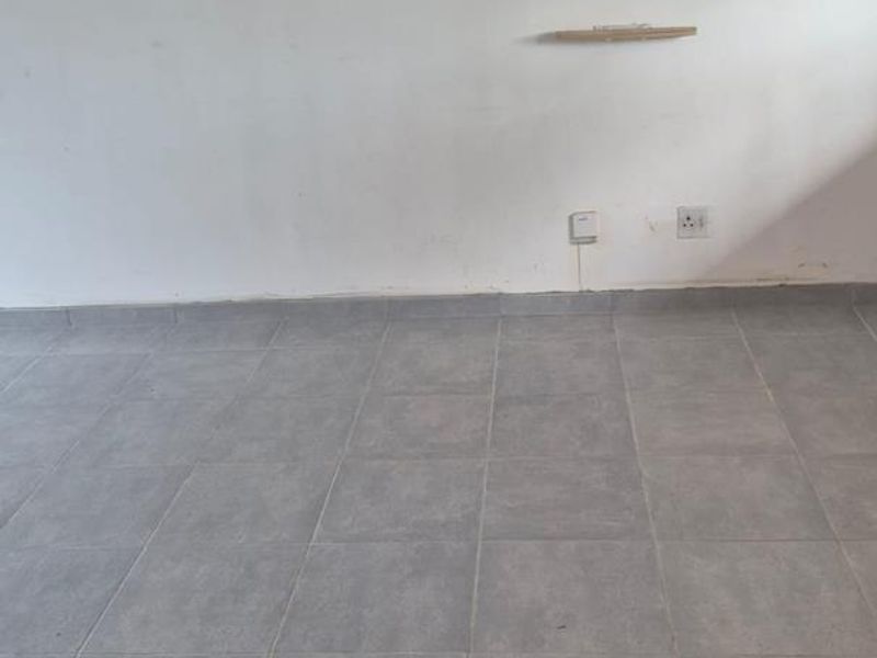 An empty living area featuring grey ceramic floor tiles, a wall-mounted shelf, and standard electrical outlets on a plain white wall.