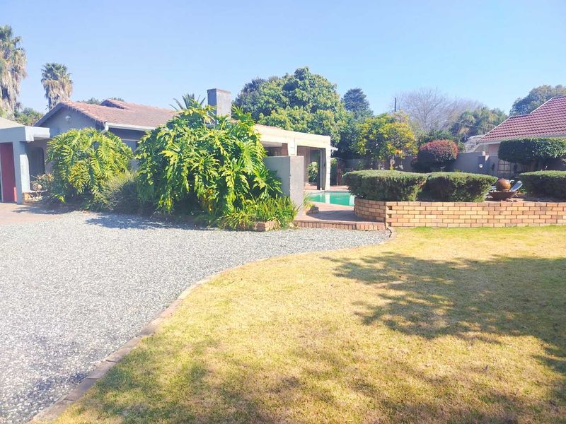 Rear exterior view featuring a swimming pool, landscaped garden with brick retaining walls, and a gravel driveway area.