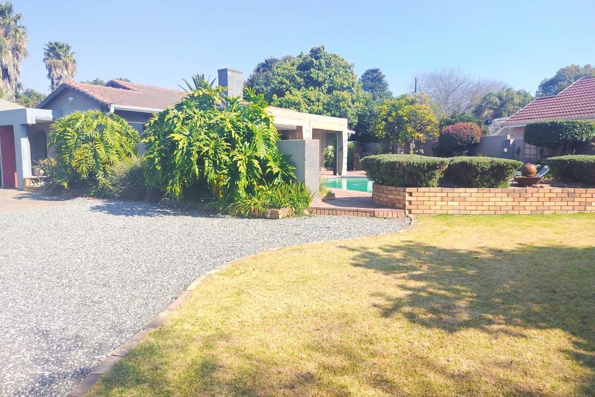 Rear exterior view featuring a swimming pool, landscaped garden with brick retaining walls, and a gravel driveway area.