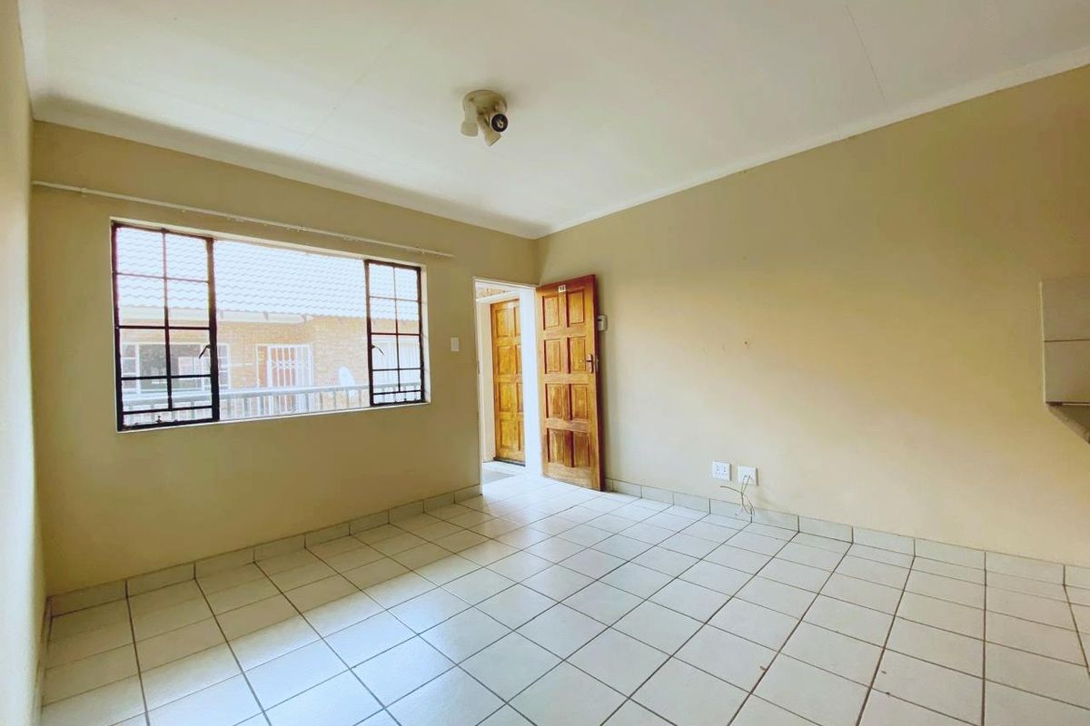 An empty living area featuring neutral tiled flooring, large windows for natural light, and a wooden door leading to an adjacent space.