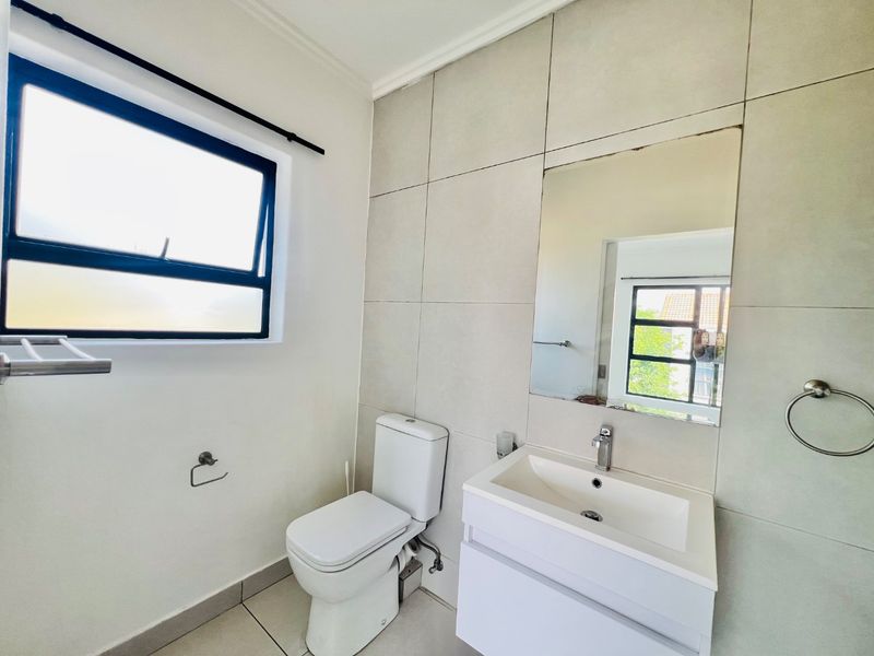 A clean, modern bathroom featuring a white vanity with basin, wall-mounted mirror, toilet, and neutral large-format wall tiles.