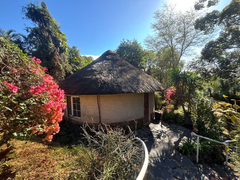 A charming circular cottage featuring a traditional thatched roof, brick walls, and leaded glass windows, set within a lush, landscaped garden with stone pathways.
