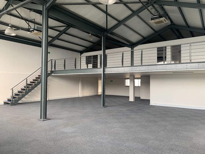 Spacious open-plan office area featuring a mezzanine level, industrial-style exposed steel structure, carpeted flooring, and integrated air conditioning units.