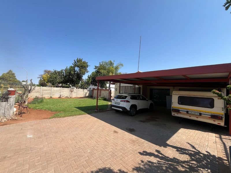 Rear exterior view featuring a paved driveway, a covered carport structure, and a well-maintained lawn area enclosed by a precast concrete boundary wall.
