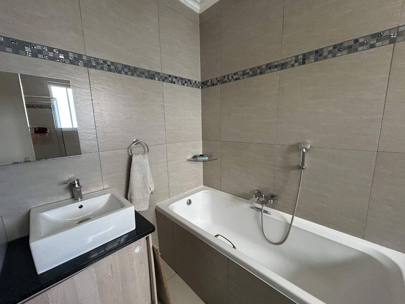 A modern bathroom featuring a vanity with basin, a bathtub with handheld shower attachment, and neutral wall tiling with decorative mosaic accents.