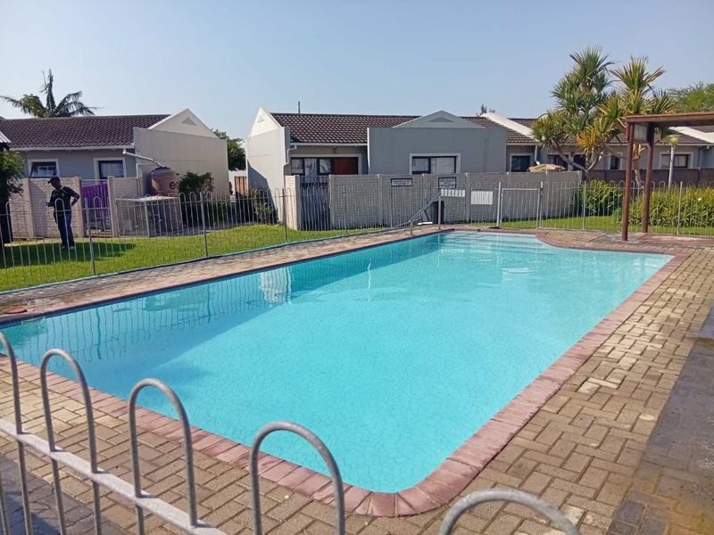 A communal swimming pool area featuring a paved deck, metal safety fencing, and a pergola, set within a well-maintained townhouse complex.