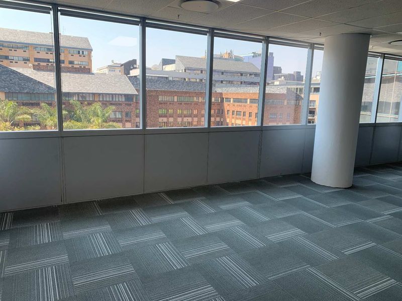 An empty, open-plan office space featuring large windows with urban views, carpet tile flooring, and a central structural column.