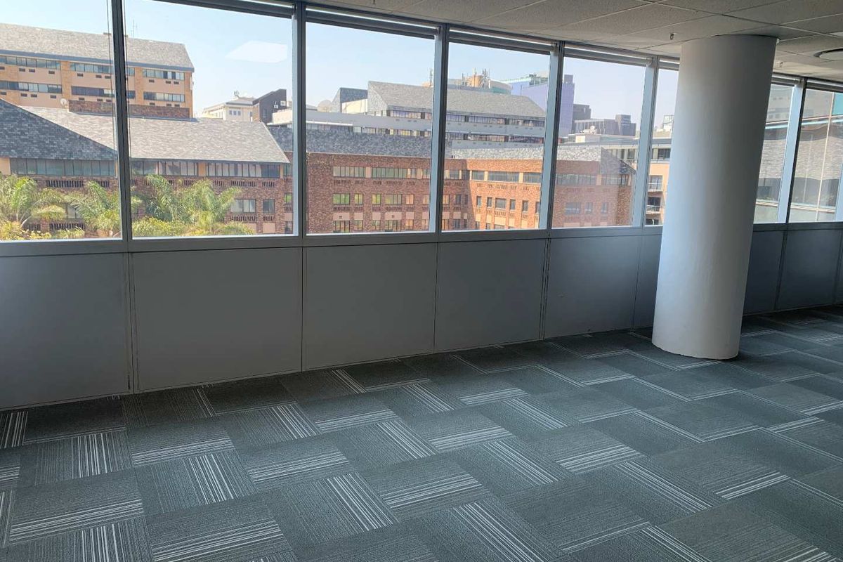 An empty, open-plan office space featuring large windows with urban views, carpet tile flooring, and a central structural column.