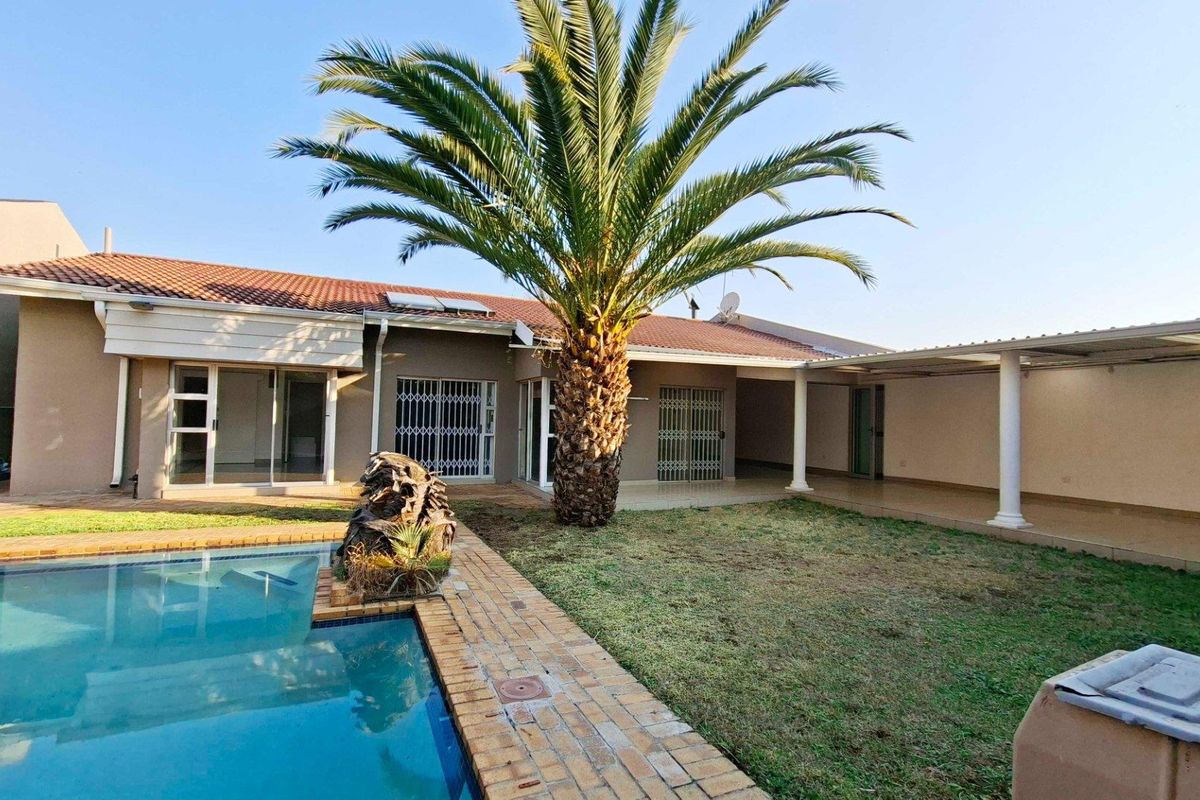 Rear exterior view featuring a swimming pool, brick-paved surround, covered patio area, and established palm tree in a private garden setting.