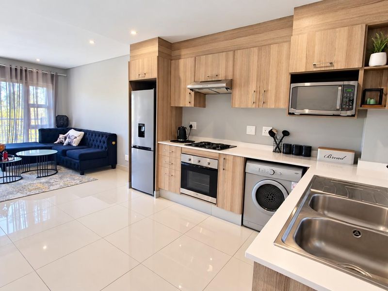 Modern open-plan kitchen and living area featuring light wood cabinetry, integrated appliances, sleek tiled flooring, and direct access to a balcony.