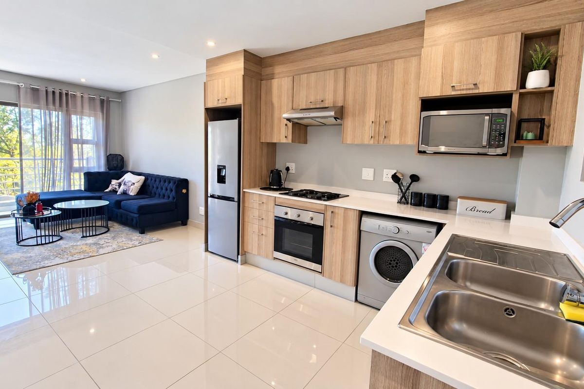 Modern open-plan kitchen and living area featuring light wood cabinetry, integrated appliances, sleek tiled flooring, and direct access to a balcony.
