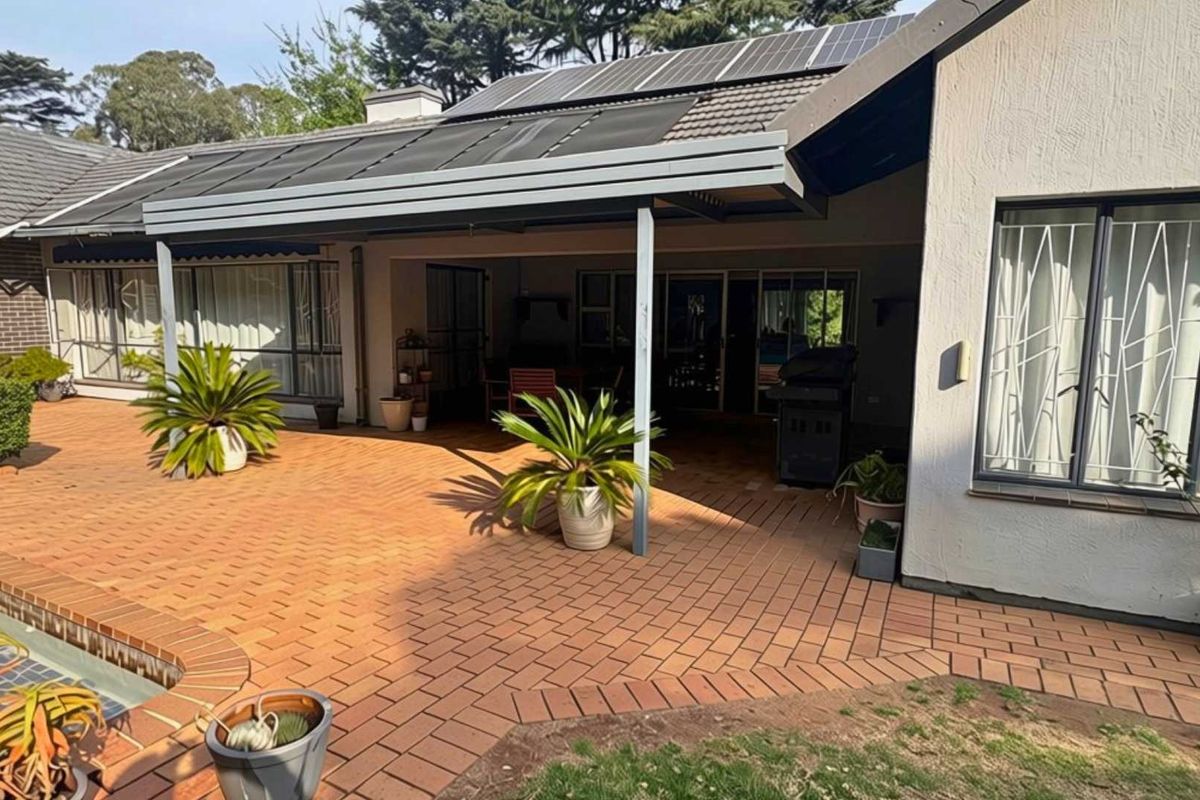A spacious covered patio area featuring brick paving, overlooking a swimming pool, with solar panels installed on the roof structure.