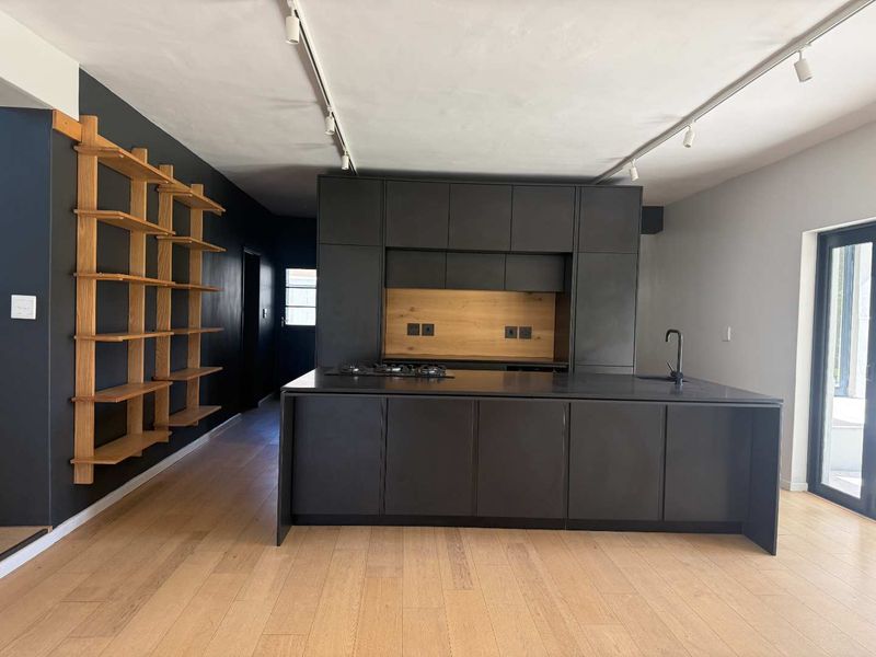 Modern open-plan kitchen featuring sleek dark cabinetry, a central island with a gas hob, integrated sink, and warm timber shelving accents.