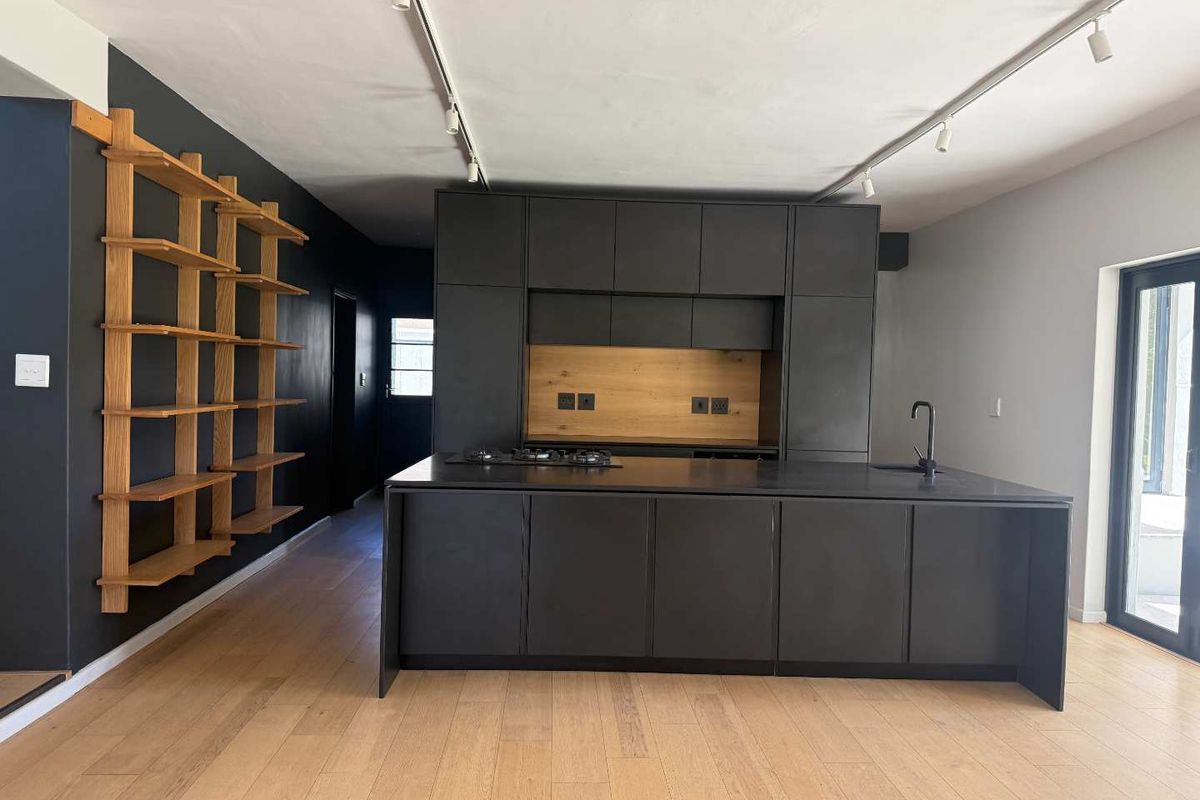 Modern open-plan kitchen featuring sleek dark cabinetry, a central island with a gas hob, integrated sink, and warm timber shelving accents.