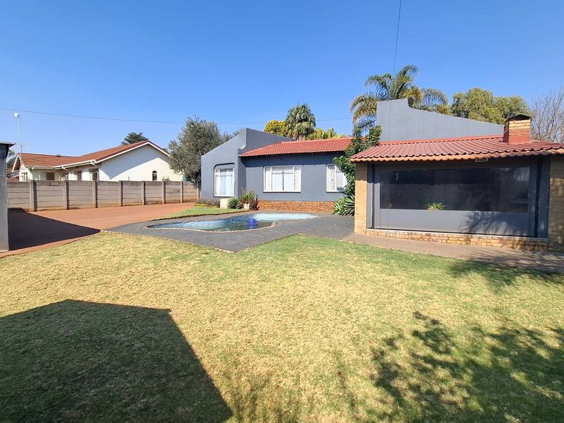 Rear exterior view featuring a swimming pool, paved patio area with outdoor blinds, and a spacious lawn enclosed by a brick boundary wall.