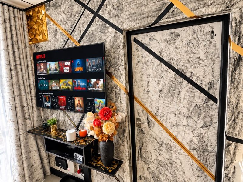 A living room area featuring a unique decorative wall finish with geometric gold and black accents, tiled flooring, and a wall-mounted television.