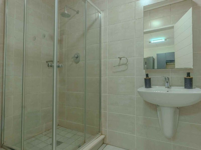 A functional bathroom featuring a glass-enclosed shower, pedestal basin, and wall-mounted mirror with neutral wall and floor tiling.