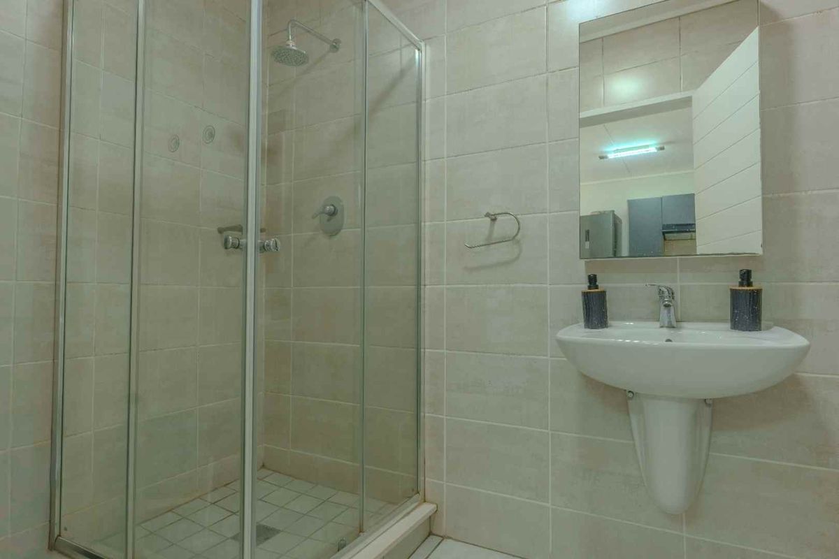 A functional bathroom featuring a glass-enclosed shower, pedestal basin, and wall-mounted mirror with neutral wall and floor tiling.