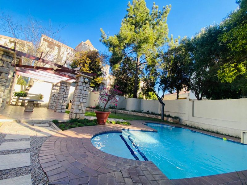 A well-maintained outdoor pool area featuring a swimming pool, stone-clad pillars, and a timber pergola, set within a private garden space.