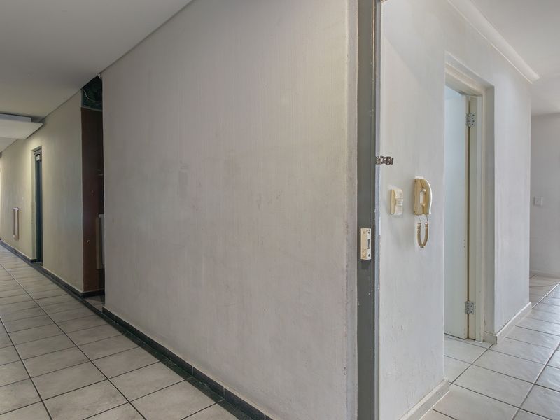 A long, tiled apartment hallway featuring recessed lighting and a wall-mounted intercom system, leading towards an open living area with natural light.