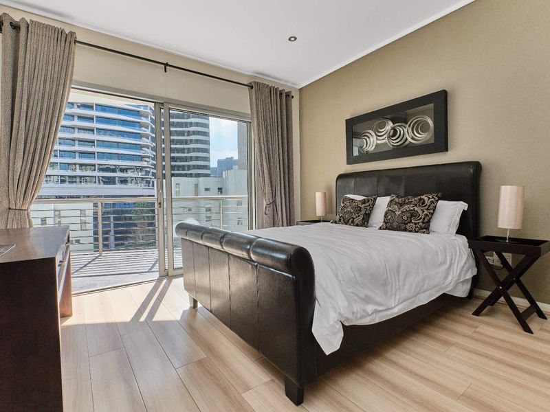 A well-maintained bedroom featuring light wood-look flooring, recessed ceiling lighting, and large sliding glass doors that open onto a private balcony.