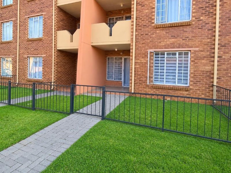 Ground floor apartment exterior featuring a brick facade, private paved walkway, and secure metal fencing surrounding a small lawn area.