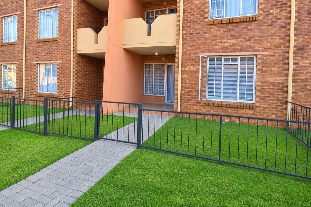 Ground floor apartment exterior featuring a brick facade, private paved walkway, and secure metal fencing surrounding a small lawn area.