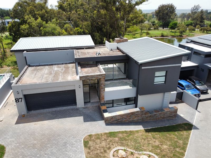 Aerial view of a modern two-story house featuring stone cladding, a paved driveway, double garage, and metal roofing, situated in a landscaped estate.
