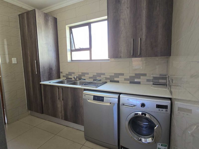A functional scullery featuring modern dark wood cabinetry, a double sink, tiled backsplash, and dedicated space for laundry and dishwashing appliances.