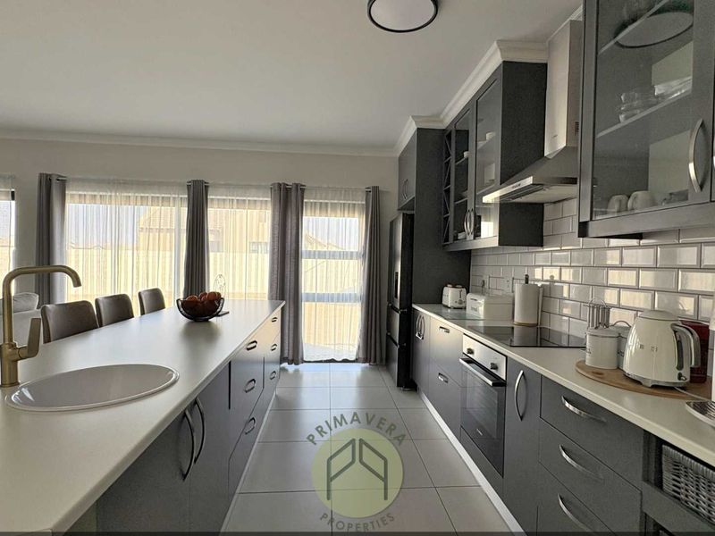 Modern open-plan kitchen featuring dark cabinetry, a large island with an undermount sink, subway tile backsplash, and integrated appliances, flowing into the living area.
