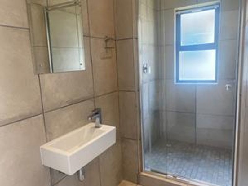 A modern bathroom featuring a wall-mounted basin, mixer tap, and a glass-enclosed shower with neutral floor-to-ceiling tiling.