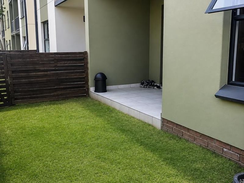 A ground floor apartment exterior featuring a private tiled patio, a small lawn area, and a timber privacy fence.
