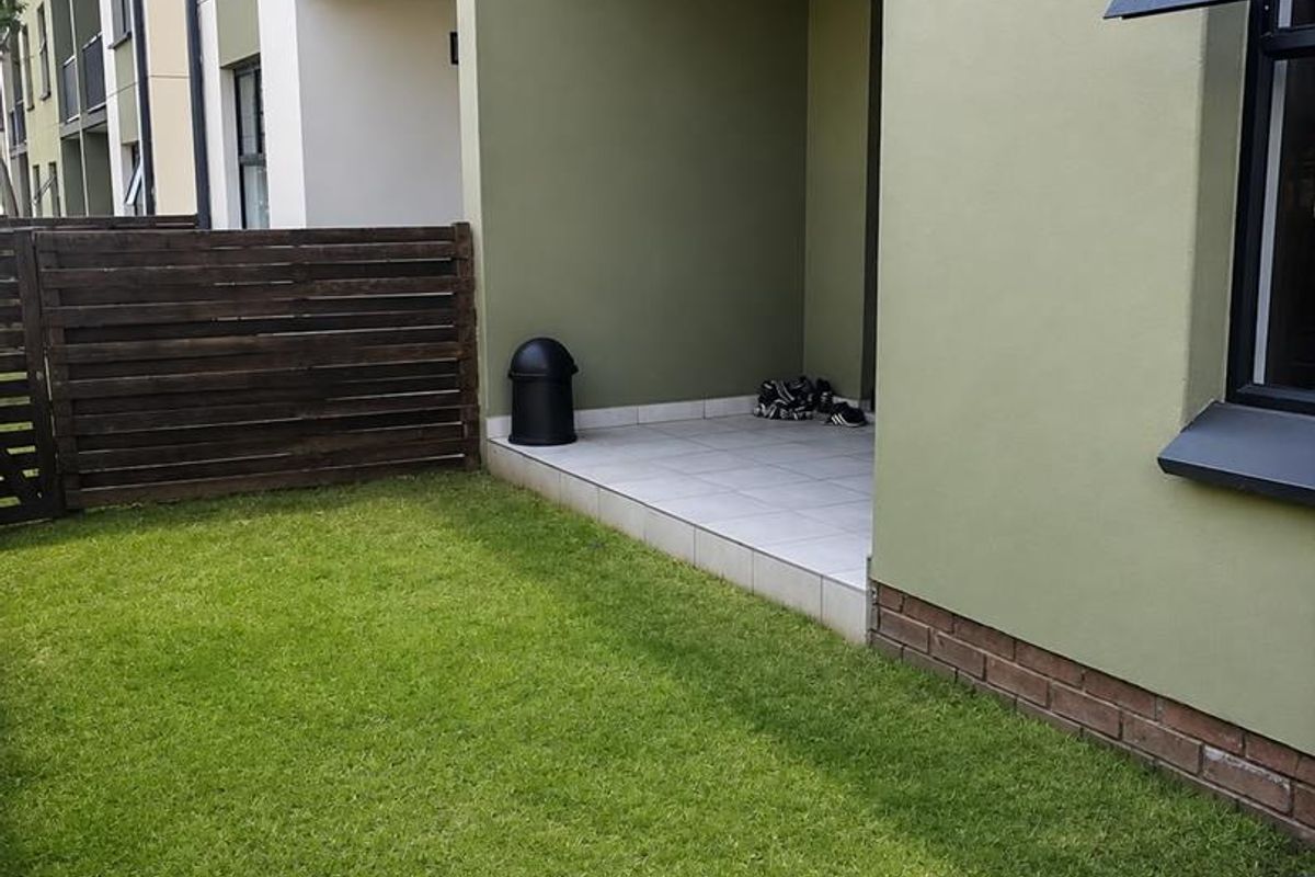 A ground floor apartment exterior featuring a private tiled patio, a small lawn area, and a timber privacy fence.