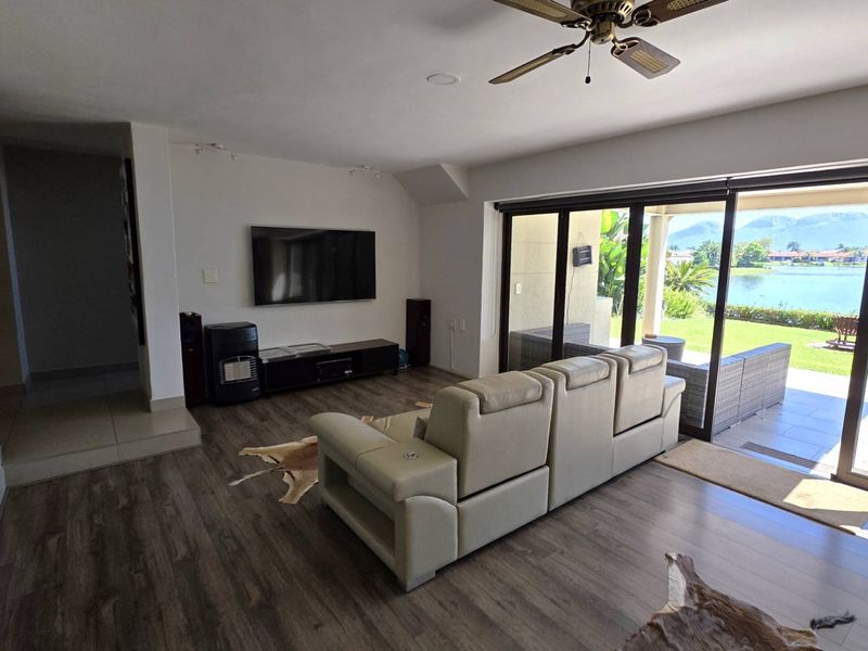 Spacious living room featuring modern laminate flooring, recessed lighting, and large sliding glass doors that open to a scenic waterfront patio and garden view.
