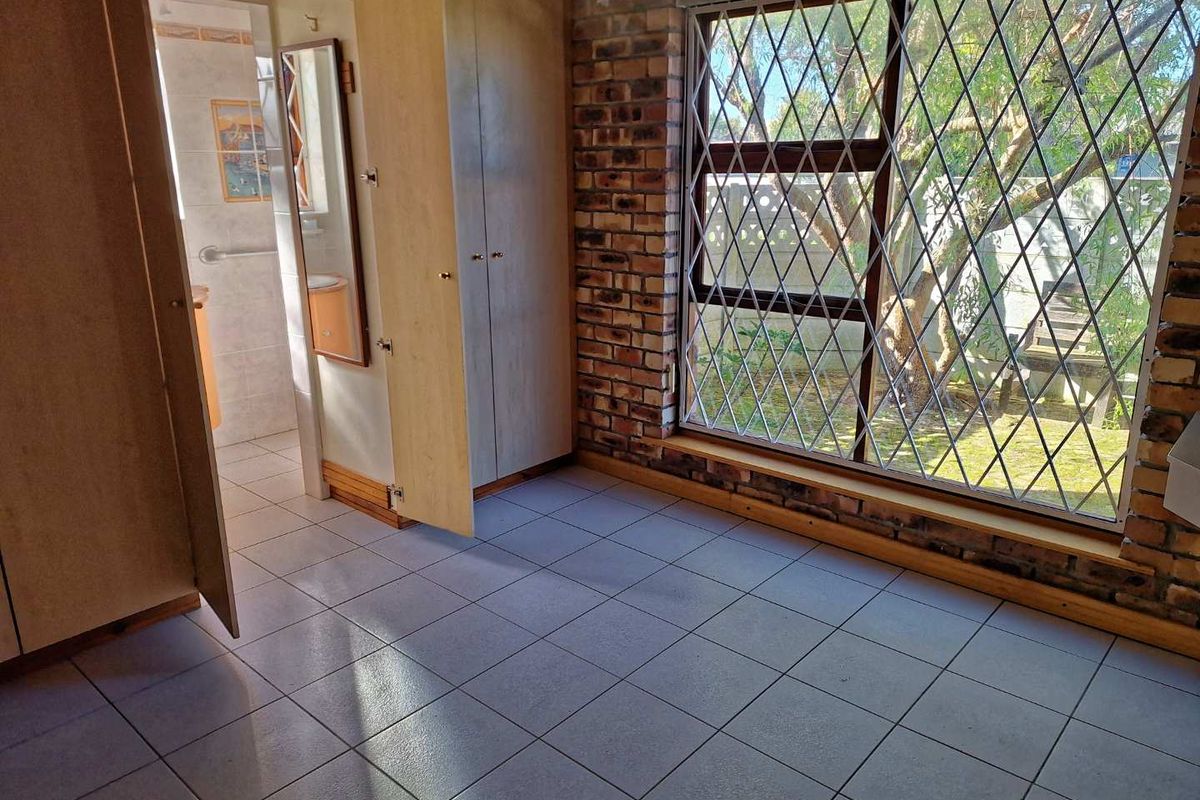 A bedroom featuring tiled flooring, built-in cupboards, and a large window with security bars, leading into an en-suite bathroom.