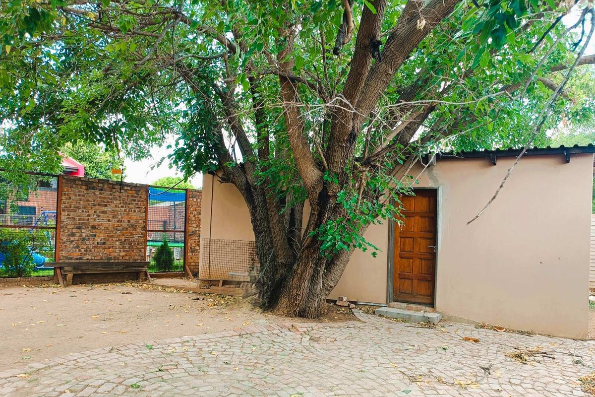 A rear exterior view featuring a paved courtyard area, a mature tree, and a solid wood door leading into a utility or storage structure.