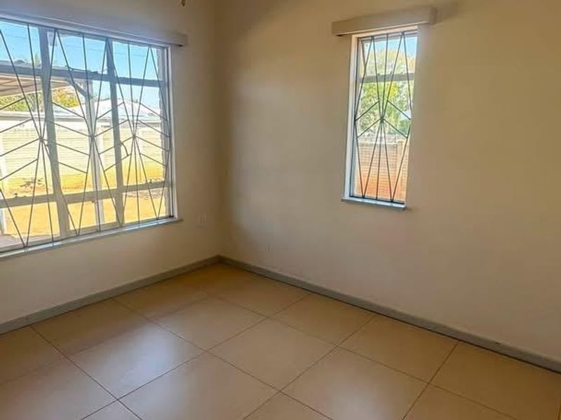 A bright, empty bedroom featuring neutral tiled flooring, a ceiling fan, and windows fitted with security bars.