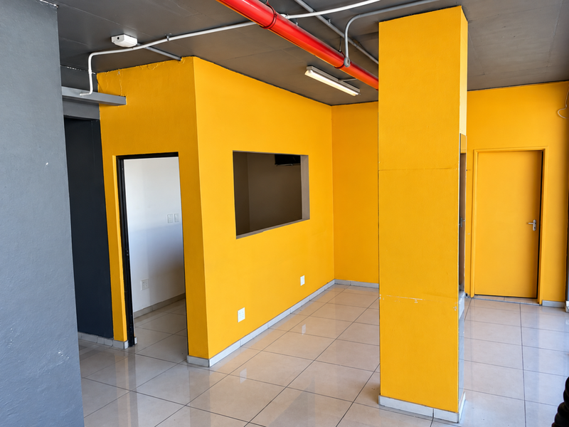 Open plan retail space featuring bright yellow walls, ceramic tiled flooring, exposed industrial ceiling piping, and a functional internal service window.