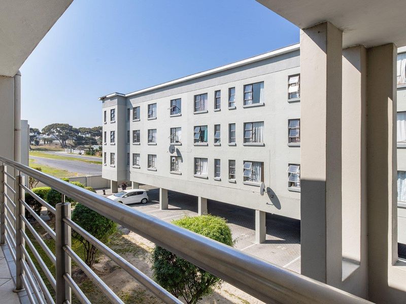 A covered balcony with tiled flooring and a stainless steel balustrade, overlooking the apartment complex grounds and parking area.