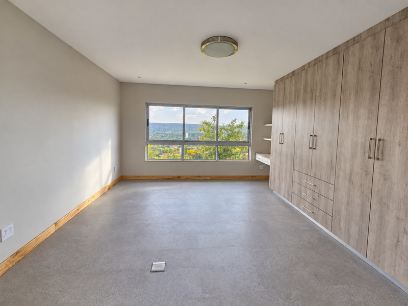 A bright, empty bedroom featuring modern wood-finish built-in cupboards, a built-in desk, tiled flooring, and large windows offering scenic views.