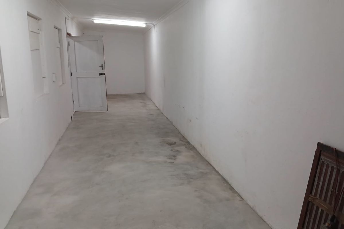 A long, empty industrial hallway featuring concrete flooring, white walls, and basic fluorescent lighting, suitable for storage or utility use.