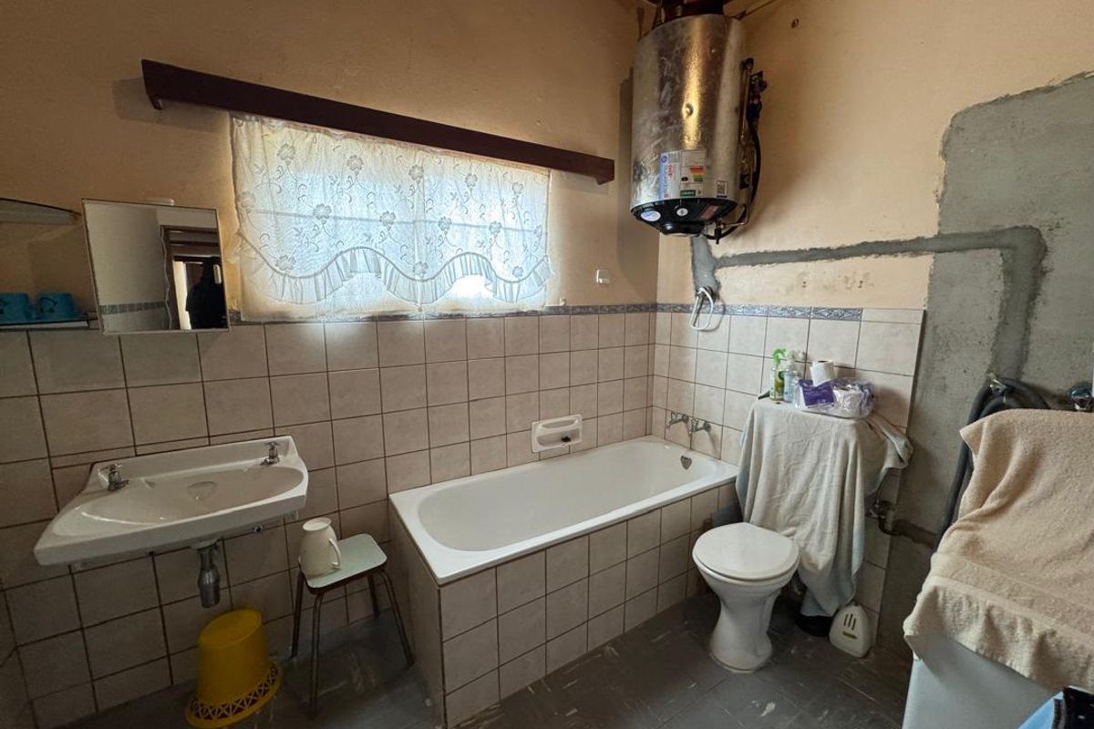 A dated bathroom featuring a standard bathtub, wall-mounted basin, and exposed electric water heater, requiring significant renovation and cosmetic updates.