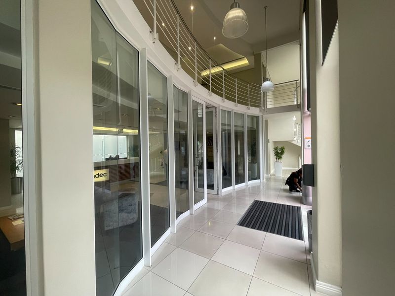 A bright office hallway featuring glass partition walls, tiled flooring, and a mezzanine level with a modern metal balustrade.