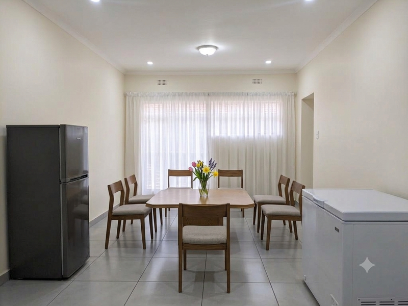 A bright and neutral dining area featuring tiled flooring, recessed downlighting, and large windows dressed with sheer curtains.