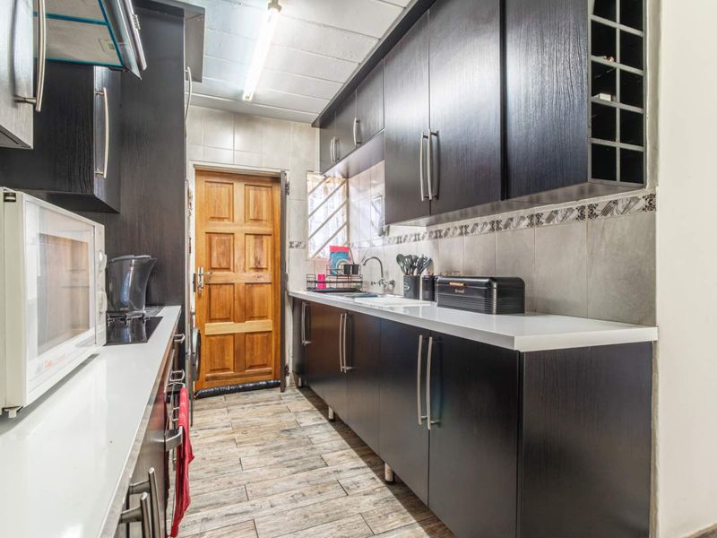 A functional galley-style kitchen featuring dark wood cabinetry, white stone countertops, tiled backsplash, and wood-look floor tiles with direct access to an exterior door.