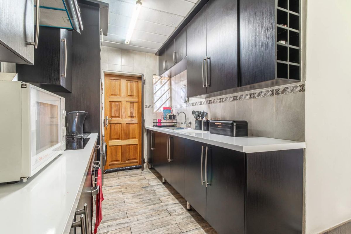 A functional galley-style kitchen featuring dark wood cabinetry, white stone countertops, tiled backsplash, and wood-look floor tiles with direct access to an exterior door.