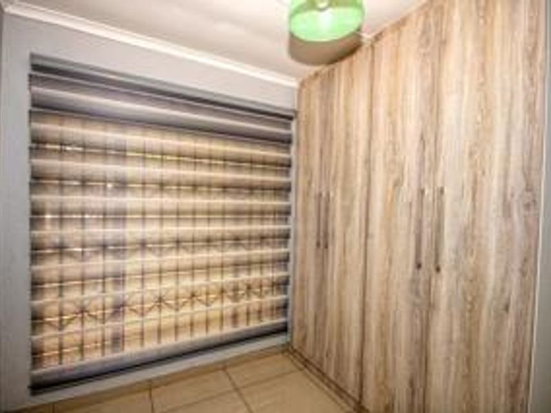 A clean bedroom featuring modern wood-finish built-in cupboards, tiled flooring, and contemporary roller blinds.