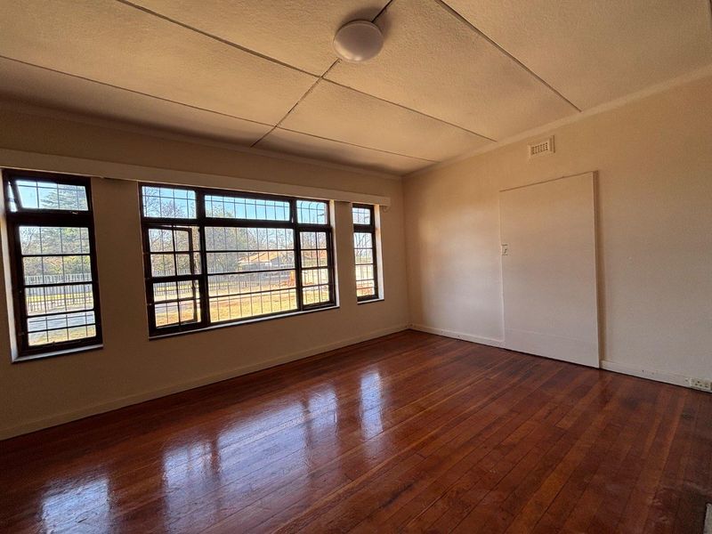 A bright, empty living room featuring polished hardwood flooring, large steel-framed windows with security bars, and a neutral interior finish.