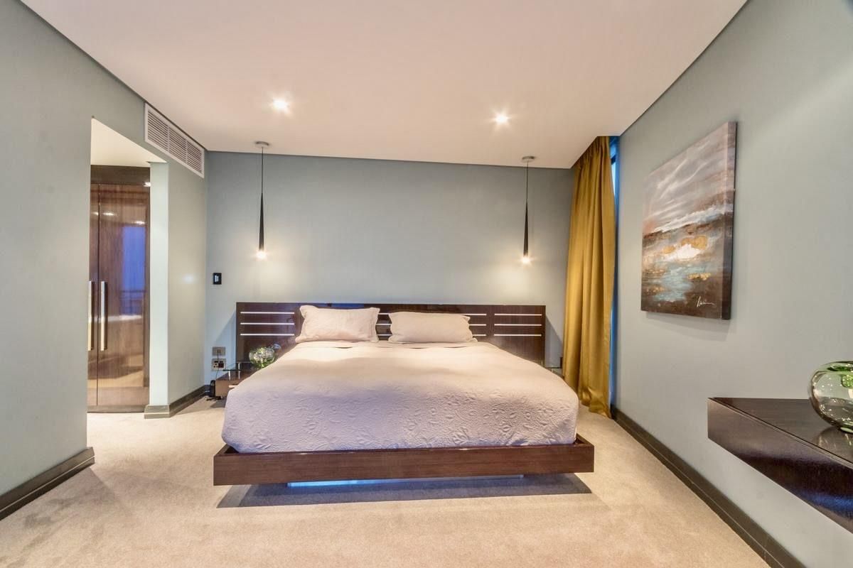 A well-appointed bedroom featuring recessed ceiling lighting, modern pendant lights, and integrated air conditioning vents, finished with neutral carpeting and soft-toned walls.