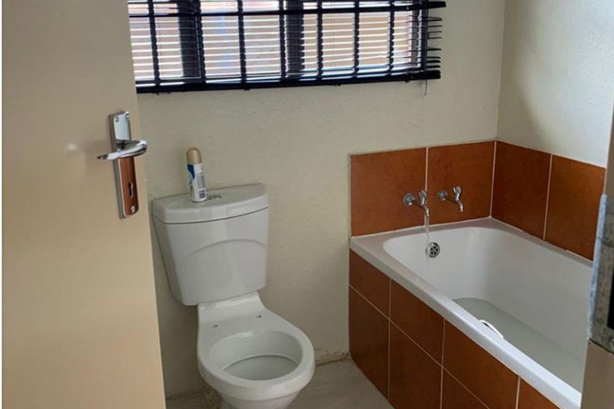 A functional bathroom featuring a built-in bathtub with terracotta-style tiling, a standard toilet, and a window fitted with security bars and blinds.